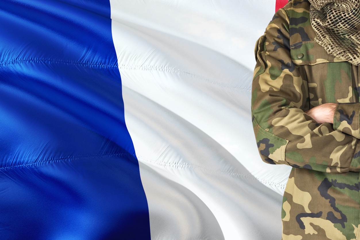 Crossed arms French soldier with national waving flag on background – France Military theme.